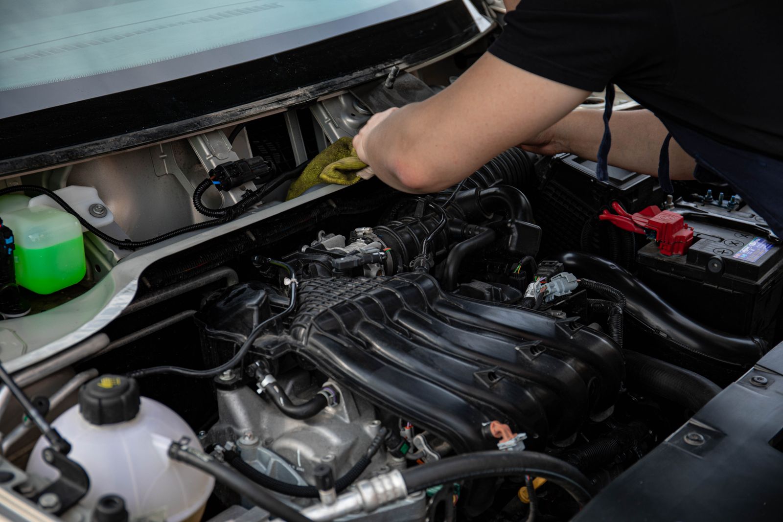 Close-up of a mechanic's arm reaching into an open engine bay with a cloth, with a coolant reservoir and battery visible