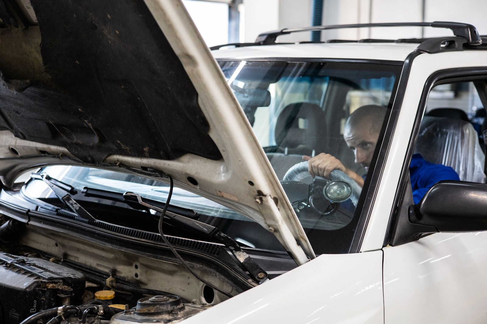 A mechanic sitting in the driver's seat of a white SUV with the bonnet open, working near the steering column area, seen through the open engine bay